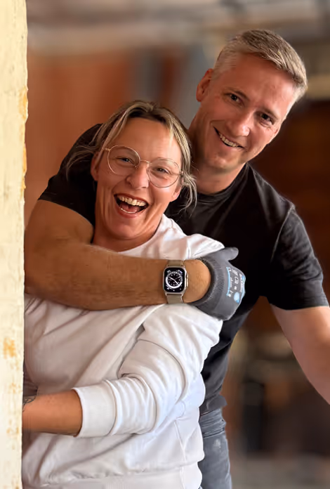 Bild Lächendes Paar, der Mann umarmt die Frau von hinten, beide tragen lässige Kleidung.